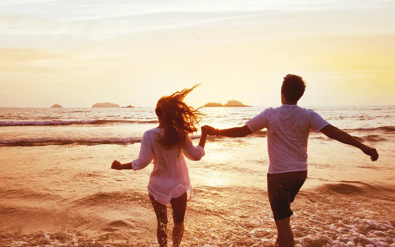 Couple-running-across-beach-towards-ocean-holding-hands